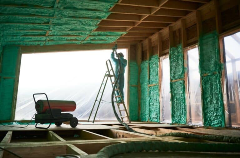 Male builder insulating wooden frame house. Man worker spraying polyurethane foam inside of future cottage, standing on ladder, using plural component gun. Construction and insulation concept.